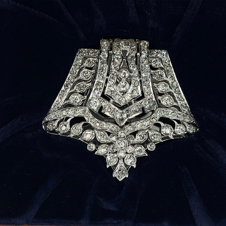 May include: A silver and diamond brooch with a geometric design. The brooch is shaped like a bell and has a large diamond in the center. The brooch is set on a dark blue velvet background.