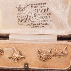 May include: Four gold-toned fox head cufflinks with small red gemstone eyes, presented in a brown rectangular hinged box. The interior lid features the printed text: CHRONOMETER MAKERS, Kendal & Dent, TO THE ADMIRALTY, THE ROYAL ITALIAN NAVY & C, 106, CHEAPSIDE, LONDON.