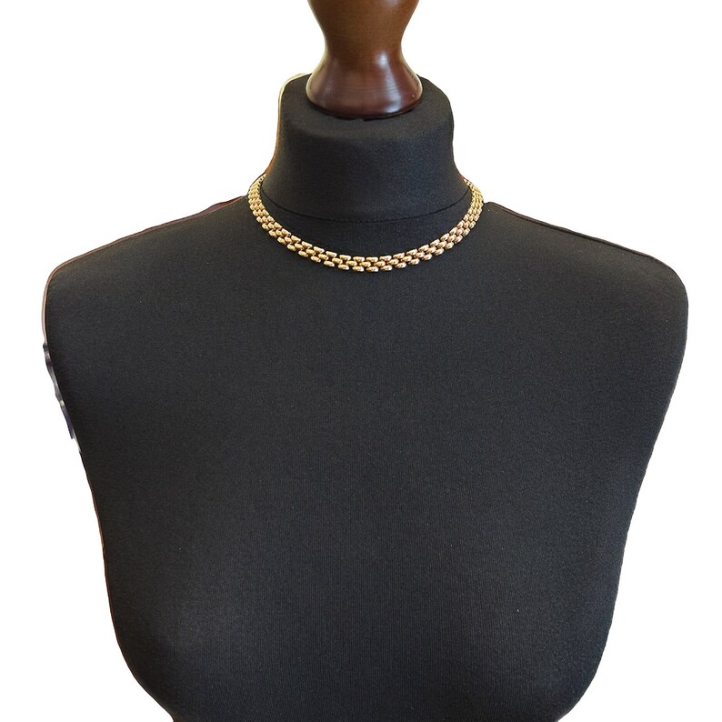 May include: A gold chain necklace on a black mannequin bust. The necklace is made of small, linked oval shapes.