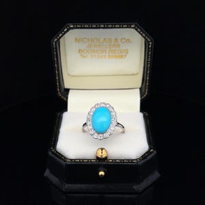 May include: A turquoise and diamond ring in a black ring box. The ring is set in silver and features a large oval turquoise stone surrounded by a halo of smaller diamonds.