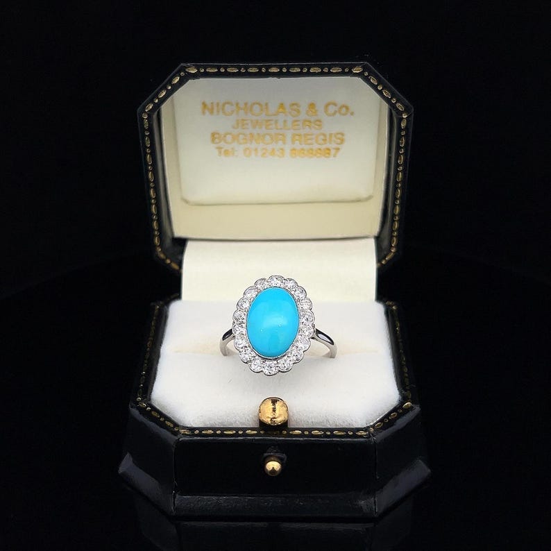 May include: A turquoise and diamond ring in a black ring box. The ring is set in silver and features a large oval turquoise stone surrounded by a halo of smaller diamonds.