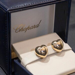 May include: A pair of polished gold heart-shaped earrings displayed in a dark blue presentation box. The interior of the box features a cream-colored velvet insert and the brand name Chopard printed in gold lettering on the back panel.
