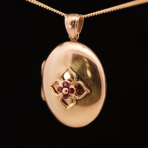 May include: A gold-colored oval locket pendant with a floral design. The pendant features a small flower with red gemstones at its center. The locket hangs from a delicate gold chain against a black background.