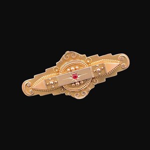 May include: A gold-toned brooch with an ornate design featuring a red gemstone in the center. The brooch has a rectangular shape with a pointed end on each side.