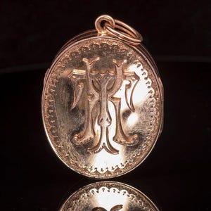 May include: An oval rose gold locket featuring an engraved monogram with the letters T and H intertwined in the centre. The locket has a decorative beaded border along the edge and includes a small circular jump ring at the top for attachment.