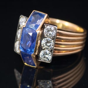 May include: A gold ring with a geometric design, featuring three round diamonds and two rectangular blue sapphires. The ring has a multi-banded gold shank. The ring is set against a black background.