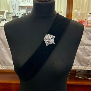 May include: A silver brooch with a geometric design, set with clear rhinestones, pinned to a black velvet sash.