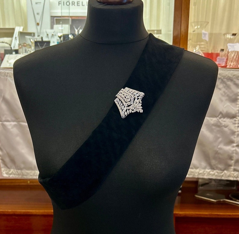 May include: A silver brooch with a geometric design, set with clear rhinestones, pinned to a black velvet sash.