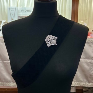 May include: A silver brooch with a geometric design featuring clear rhinestones. The brooch is pinned to a black velvet sash.