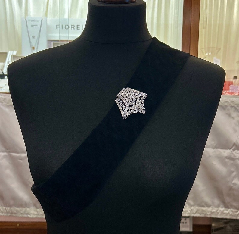 May include: A silver brooch with a geometric design featuring clear rhinestones. The brooch is pinned to a black velvet sash.
