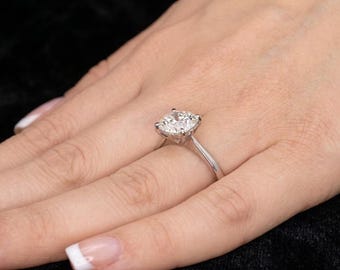 May include: A silver-toned ring featuring a single, large, round-cut clear gemstone held in a four-prong setting on a smooth, polished metal band. The ring is worn on a finger, showcasing the clarity and brilliance of the central stone.