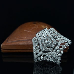 May include: A silver and diamond brooch with a geometric design. The brooch is set with small, round diamonds and has a delicate, intricate design.
