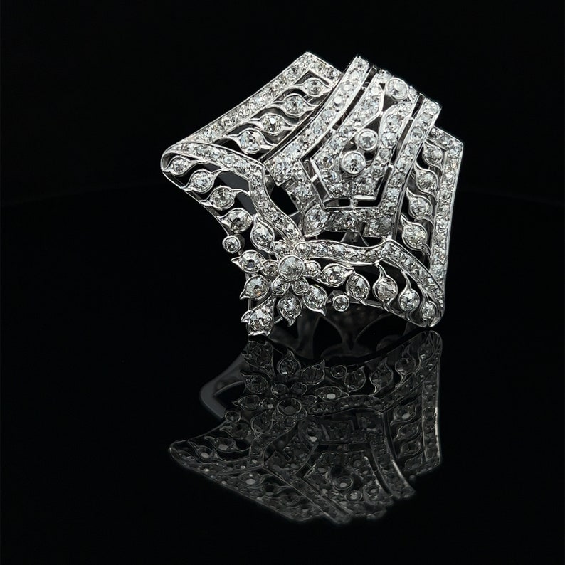 May include: A silver and diamond brooch with a geometric design. The brooch is set with small diamonds and has a floral motif in the center.
