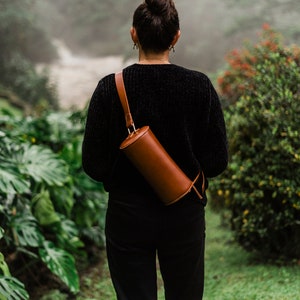May include: A person wearing a black sweater and black pants with a brown leather cylindrical bag slung over their shoulder.
