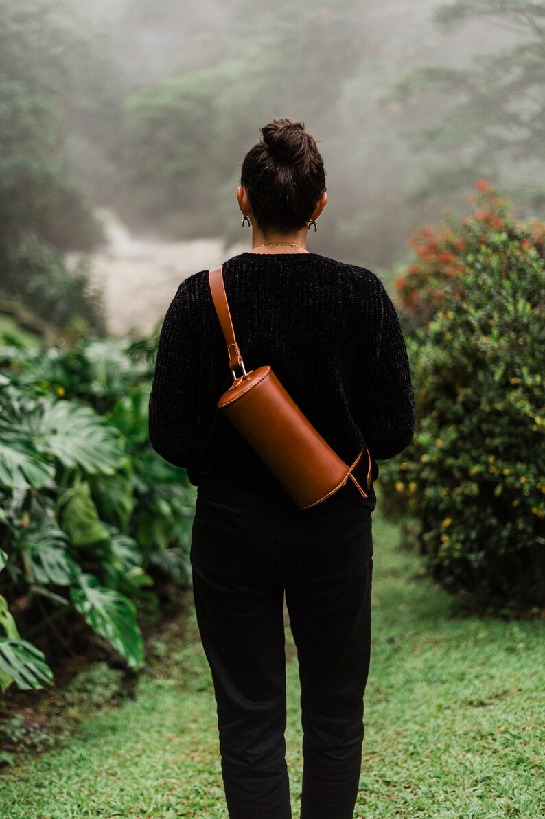 May include: A person wearing a black sweater and black pants with a brown leather cylindrical bag slung over their shoulder.