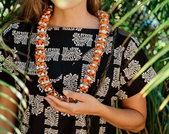 May include: A woman wearing a black and white patterned shirt with a floral lei around her neck. The lei is made of orange and white flowers.