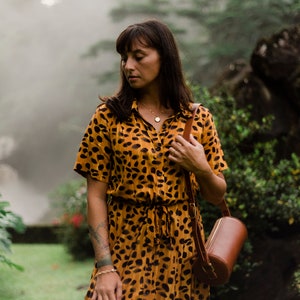 May include: A woman wearing a yellow and black animal print dress with a brown leather cylindrical bag slung over her shoulder. She is standing in a lush green forest setting.