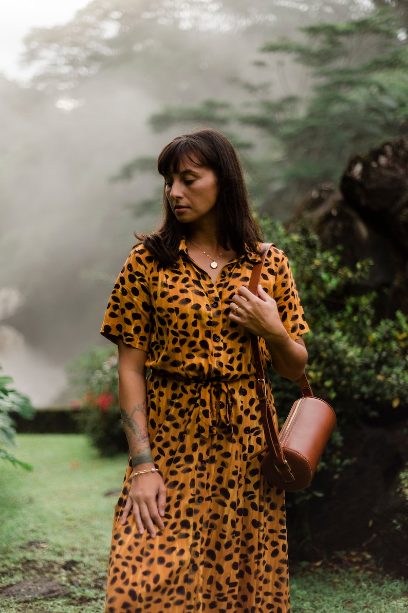 May include: A woman wearing a yellow and black animal print dress with a brown leather cylindrical bag slung over her shoulder. She is standing in a lush green forest setting.