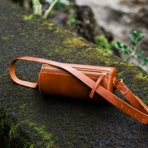 May include: A brown leather cylindrical crossbody bag with a zipper closure and an adjustable strap. The bag is lying on a mossy rock.