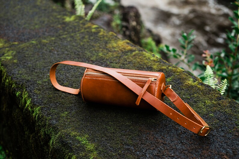 May include: A brown leather cylindrical crossbody bag with a zipper closure and an adjustable strap. The bag is lying on a mossy rock.