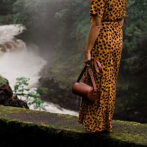 May include: A woman in a yellow and brown animal print dress stands on a mossy ledge overlooking a waterfall. She is holding a brown leather cylindrical bag with a strap.