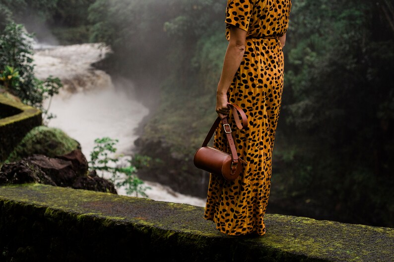 May include: A woman in a yellow and brown animal print dress stands on a mossy ledge overlooking a waterfall. She is holding a brown leather cylindrical bag with a strap.
