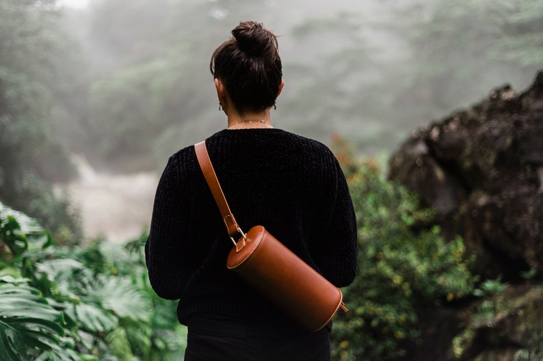 May include: A person wearing a black sweater and a brown leather cylindrical crossbody bag. The bag has a gold buckle and a long strap.