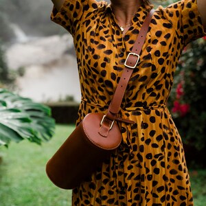 May include: A woman wearing a yellow and black animal print dress with a brown leather cylindrical bag slung over her shoulder.