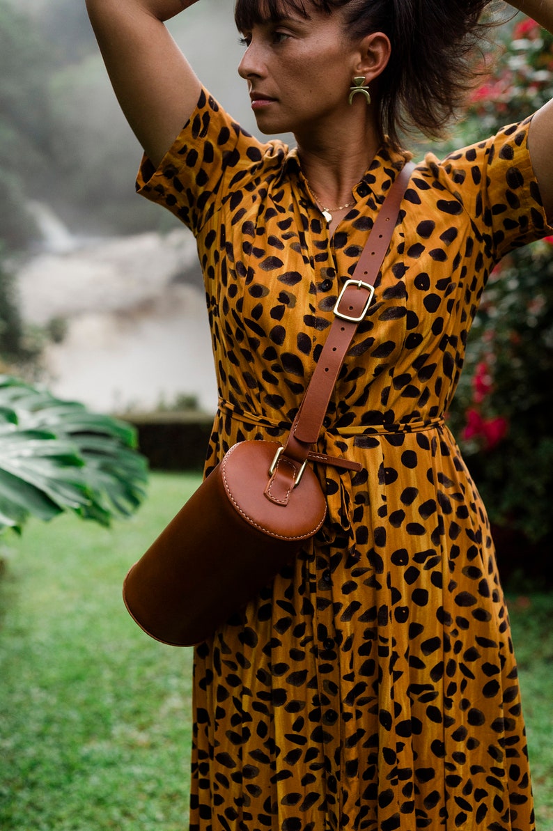 May include: A woman wearing a yellow and black animal print dress with a brown leather cylindrical bag slung over her shoulder.