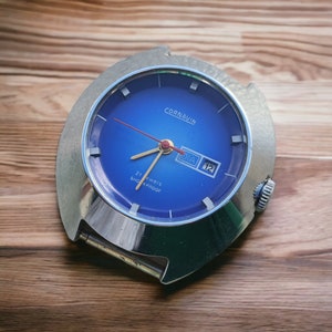 May include: A vintage Cornavin wristwatch with a vibrant blue face, gold hands, and a red second hand. The watch features a date display and is encased in a silver-toned metal frame. The text "21 jewels" and "Shockproof" are visible on the watch face.