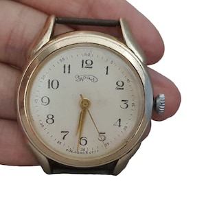 May include: A vintage gold-toned wristwatch with a white face and black leather band. The watch face features black numerals, gold hands, and the Cyrillic word "Урал". The watch has a small crown and the text "Made in USSR".