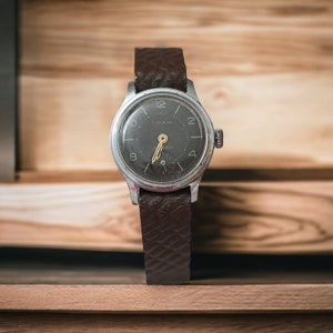 May include: Vintage wristwatch with a dark gray face, silver casing, and brown leather band. The watch face features the word "YPAH" and gold-colored hands and markers. The watch is displayed against a wooden background.