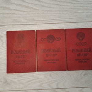 May include: Three red military documents are displayed on a light wood surface. The documents have Cyrillic text and the emblem of the USSR. The text "Military ID" is visible on each document. The documents are slightly worn.