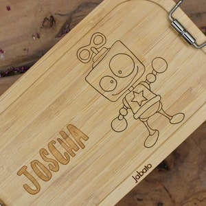 Könnte beinhalten: Eine Lunchbox aus Holz mit einem Comic-Roboter-Design und dem Namen "JOSCHA" auf der Vorderseite. Die Lunchbox hat einen silbernen Verschluss und der Markenname "Jabato" ist auf der Unterseite aufgedruckt.