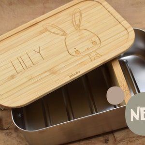 Könnte beinhalten: Eine Lunchbox aus Holz mit einem Edelstahl-Innenraum. Der Deckel ist mit einem Hasendesign und dem Namen "Lilly" graviert. Die Lunchbox ist mit "Jabato" gekennzeichnet und hat einen herausnehmbaren Trenner.