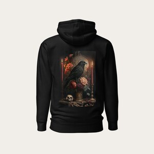 Pode incluir: Camisola de capuz preta com uma estampa de um corvo preto empoleirado em um vaso de flores com um crânio na base. As flores são vermelhas, laranja e rosa. Há duas velas acesas de cada lado do vaso.