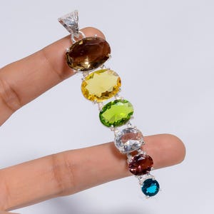 May include: A vertical pendant featuring six oval-cut gemstones in a row, including brown, yellow, lime green, clear, reddish-purple, and blue stones. The stones are set in silver metal with a decorative, textured bail at the top for hanging.