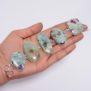 Könnte beinhalten: Ein silbernes Armband mit fünf ovalen, grün-rosa Edelsteinen. Das Armband hat einen Karabinerverschluss.