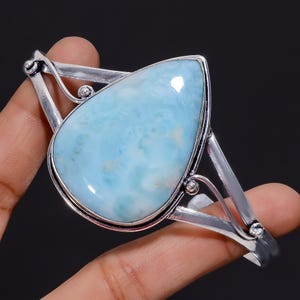 Könnte beinhalten: Ein silbernes Armreif-Armband mit einem großen, tropfenförmigen blauen Edelstein. Der Edelstein hat eine wolkig-marmorierte Optik.