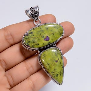 Puede incluir: Un colgante de plata con dos piedras pulidas de color verde lima con motas oscuras y pequeñas inclusiones púrpuras. Las piedras están engastadas en un marco de plata decorativo con una reasa de filigrana en la parte superior.