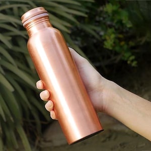 May include: A copper-colored water bottle with a screw-on cap is held in a hand. The bottle has a cylindrical shape and a shiny, metallic finish. The background is blurred, with green foliage visible.
