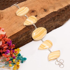 May include: A silver bracelet featuring five oval and teardrop-shaped, light yellow gemstones with brown veining. The bracelet is displayed on a piece of wood with colorful flowers in the background.
