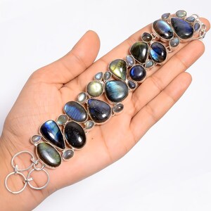 May include: A silver bracelet with a collection of iridescent labradorite stones in different shapes. The stones show blues, greens, and yellows. The bracelet is held in a hand, highlighting its design and craftsmanship. The bracelet is a piece of jewellery.