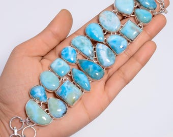 May include: A multi-stone bracelet featuring numerous polished, sky-blue Larimar gemstones set in silver. The stones vary in shape, including teardrops, ovals, and squares, arranged in a wide, linked pattern with a circular silver clasp at the end.