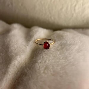May include: A delicate gold ring featuring a vibrant, oval-shaped red gemstone. The ring's slender band and the gemstone's rich color create a simple yet elegant design. The ring is set against a soft, white background.