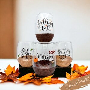May include: Four stemless wine glasses with fall-themed designs. The glasses are filled with brown and gold glitter. The designs include "Falling for Fall", "Welcome Fall", "Fall is in the air", and "Nuts for Fall".