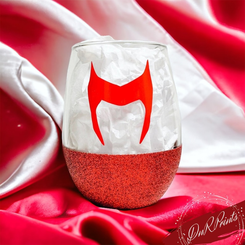 Op de afbeelding: Een rood glitter wijnglas met een rode Scarlet Witch logo sticker. Het glas staat op een rode en witte stof.
