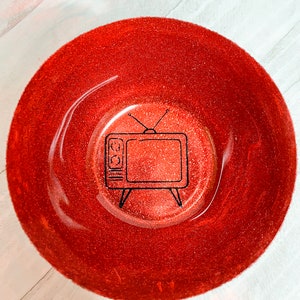 Op de afbeelding: Een rode glitterkom met een zwarte omtrek van een vintage televisie in het midden.