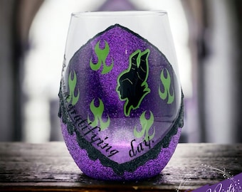 May include: Purple wine glass with a black silhouette of Maleficent from Disney's Sleeping Beauty. The glass has green flames and glitter. The text on the glass reads "It's a terrifying day."