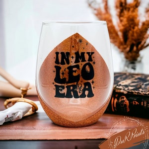May include: A clear glass wine glass with a gold glitter bottom. The glass has a black vinyl decal that says "In My Leo Era" with stars around the text.